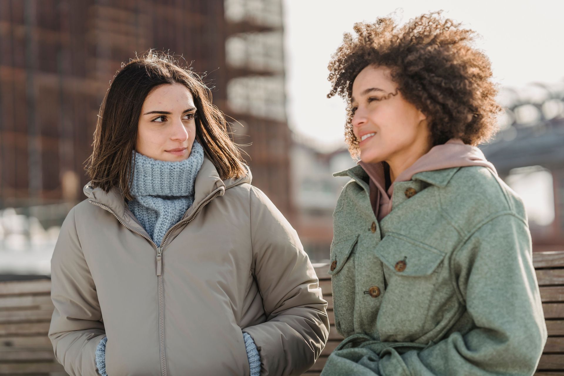 Two people in winter jackets chatting outdoors, one smiling, with a blurred city background