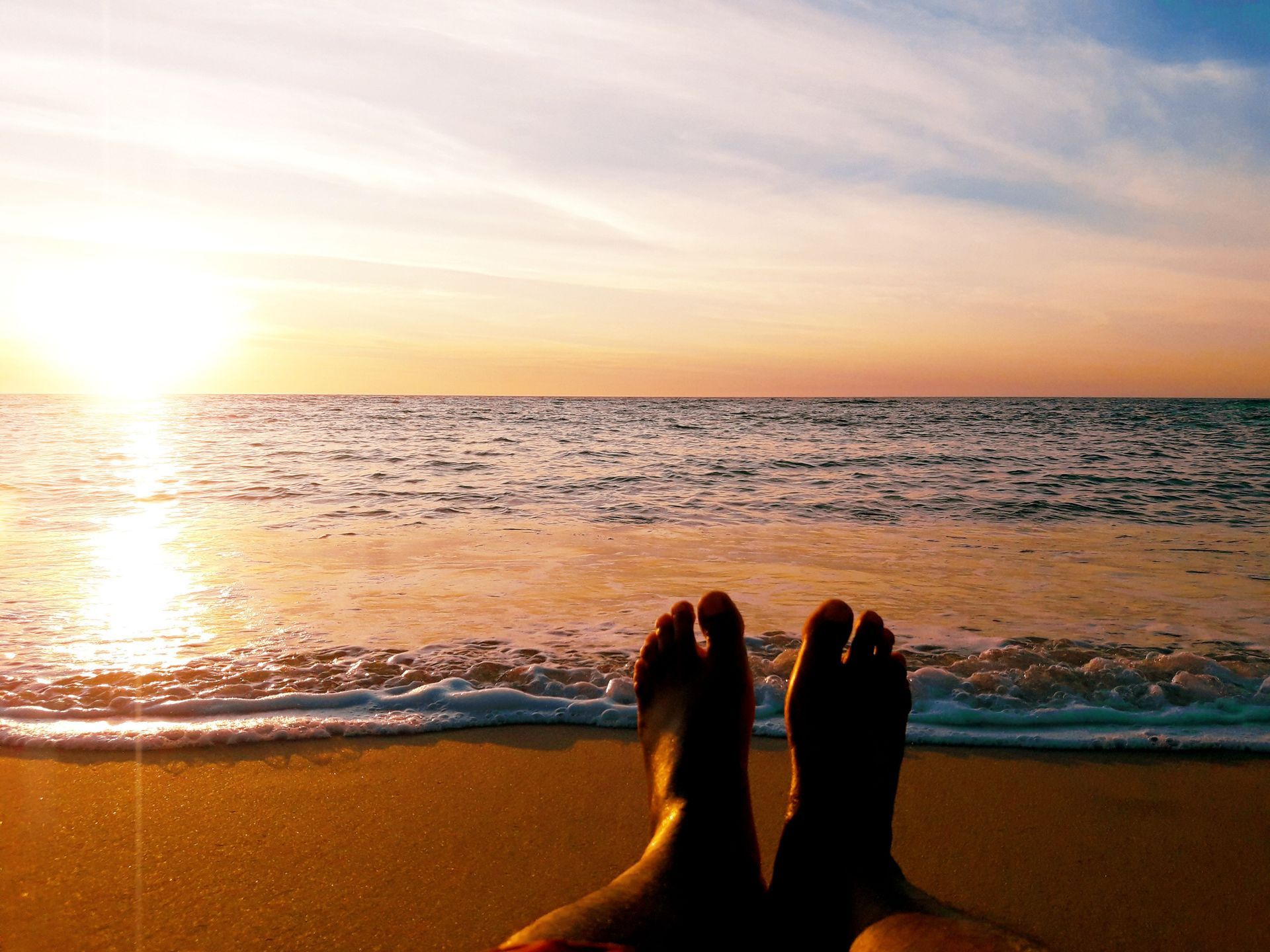 Feet in the sand facing a sunset over the ocean, with waves and a warm orange sky.