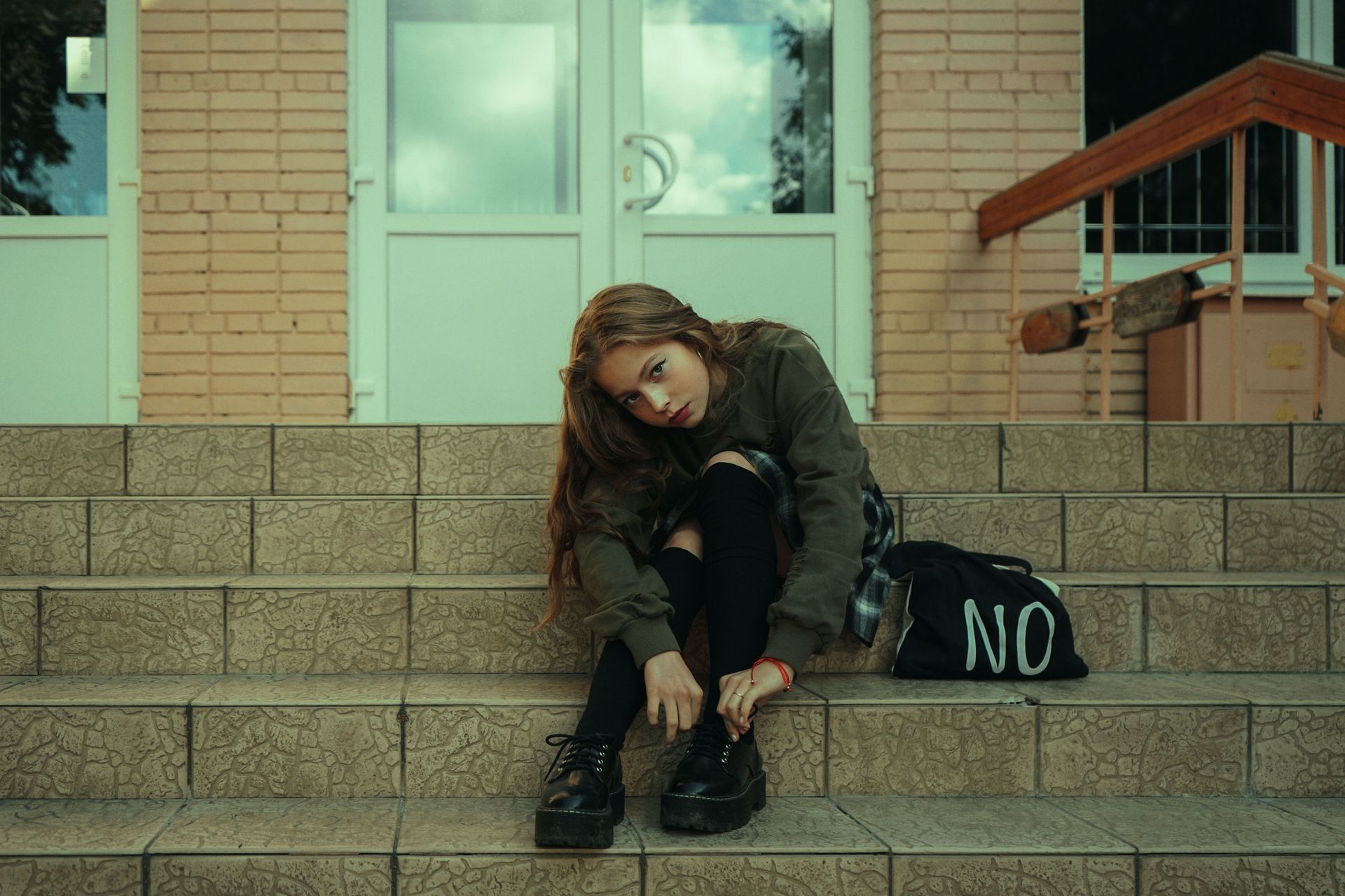 Person sits on steps outside a building, head down, beside a black bag with “NO” on it.