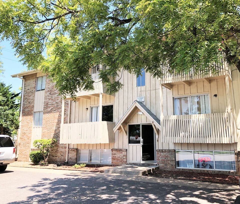 Apartment building with light wood siding and balconies, shaded by a large tree.