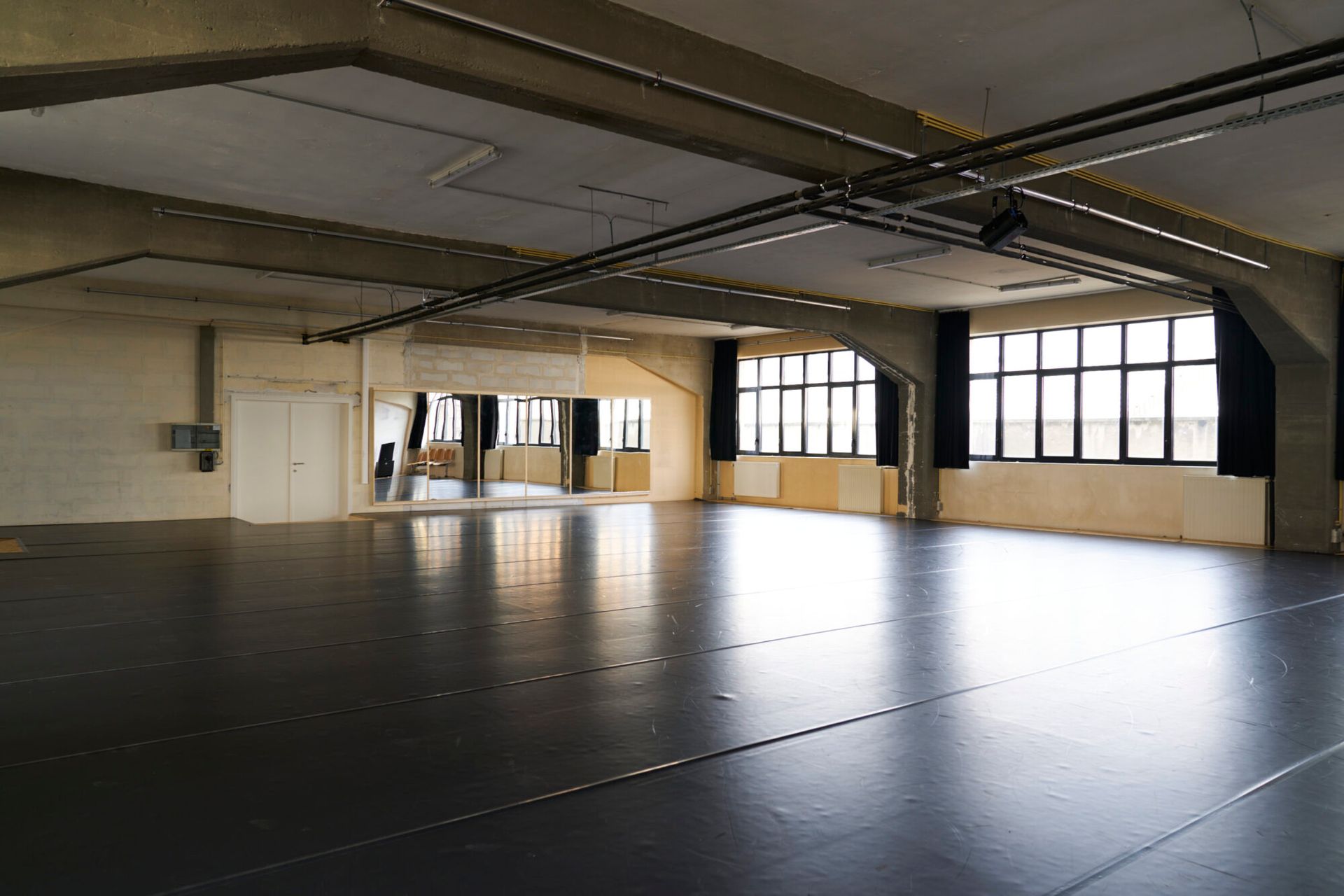 Een lege dansstudio met een zwarte vloer, spiegels, ramen en een betonnen plafond.