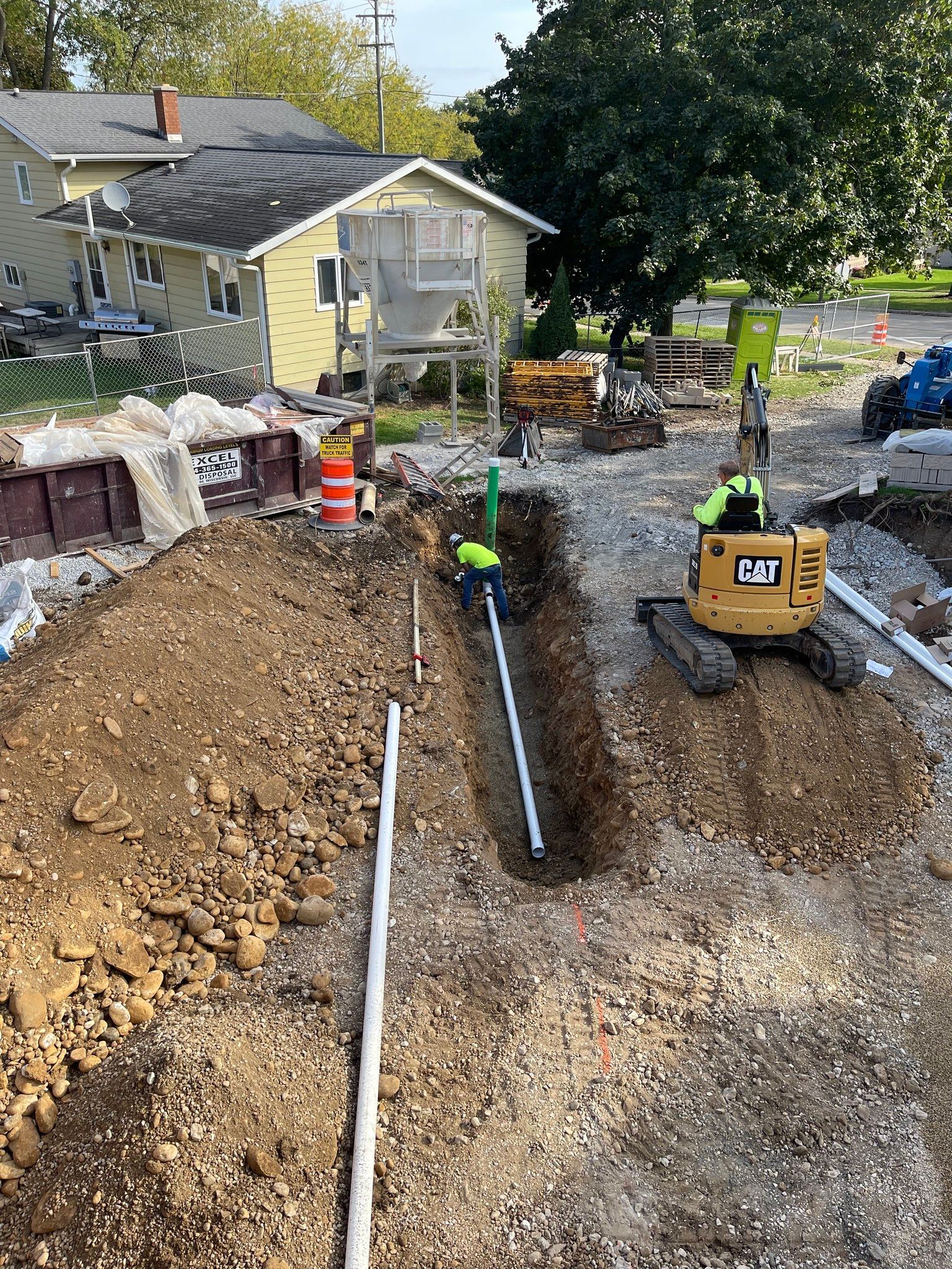 Backyard trench excavation with yellow excavator, exposed pipes, and a house in the background