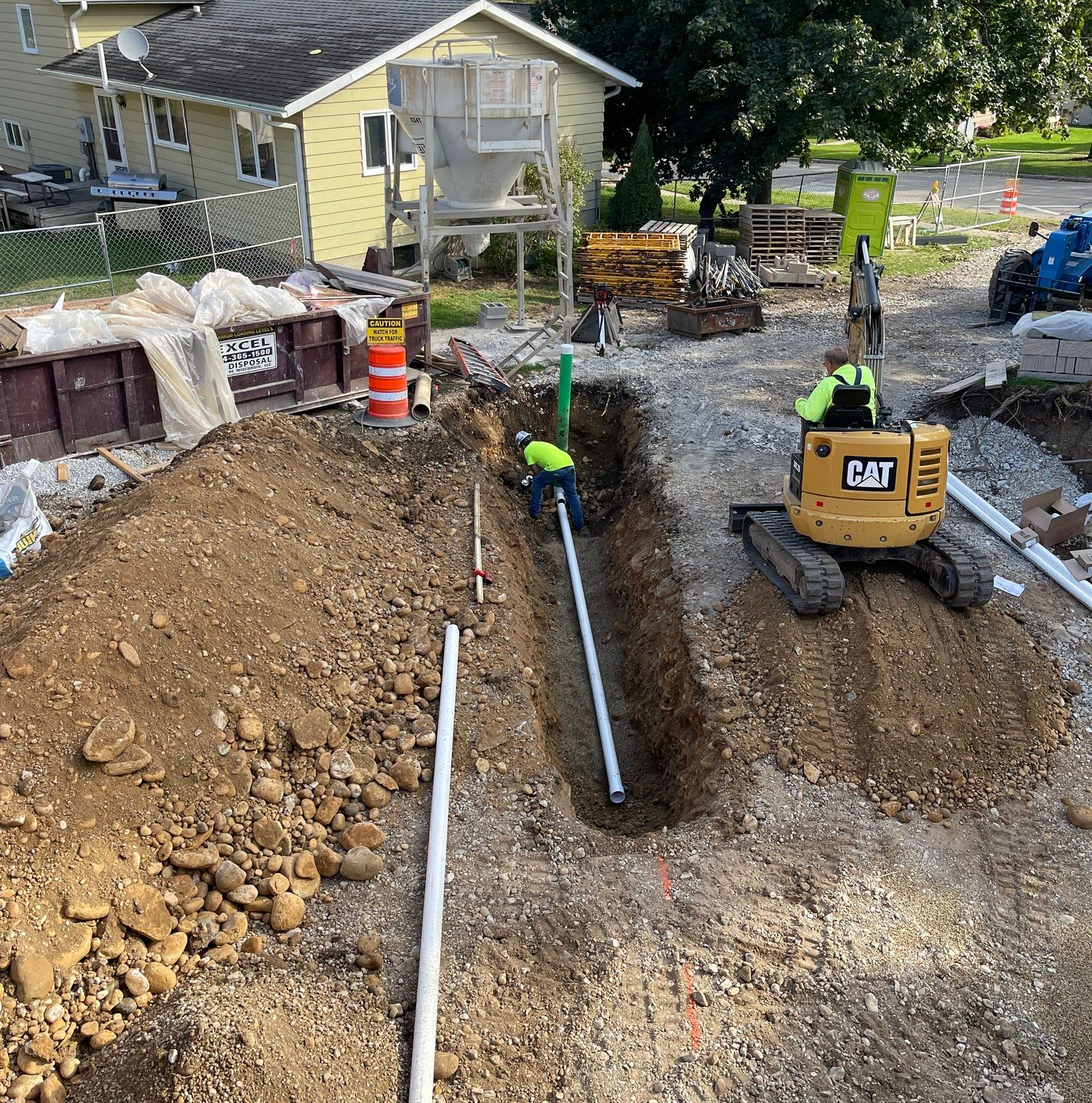 Construction workers laying white pipes in a trench beside a yellow CAT excavator at a house site