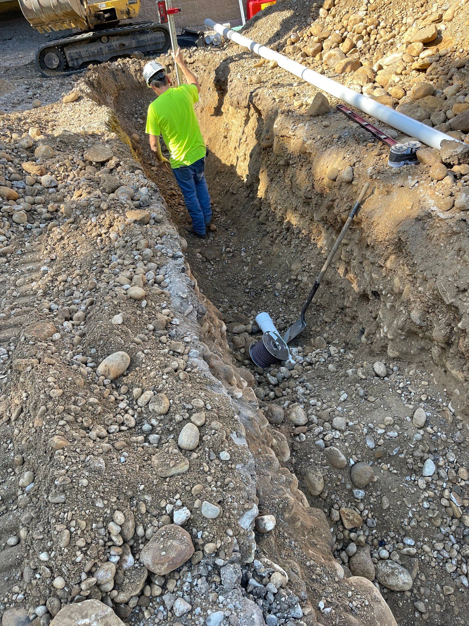 Worker in a neon shirt stands in a deep utility trench with pipes and gravel on a construction site.