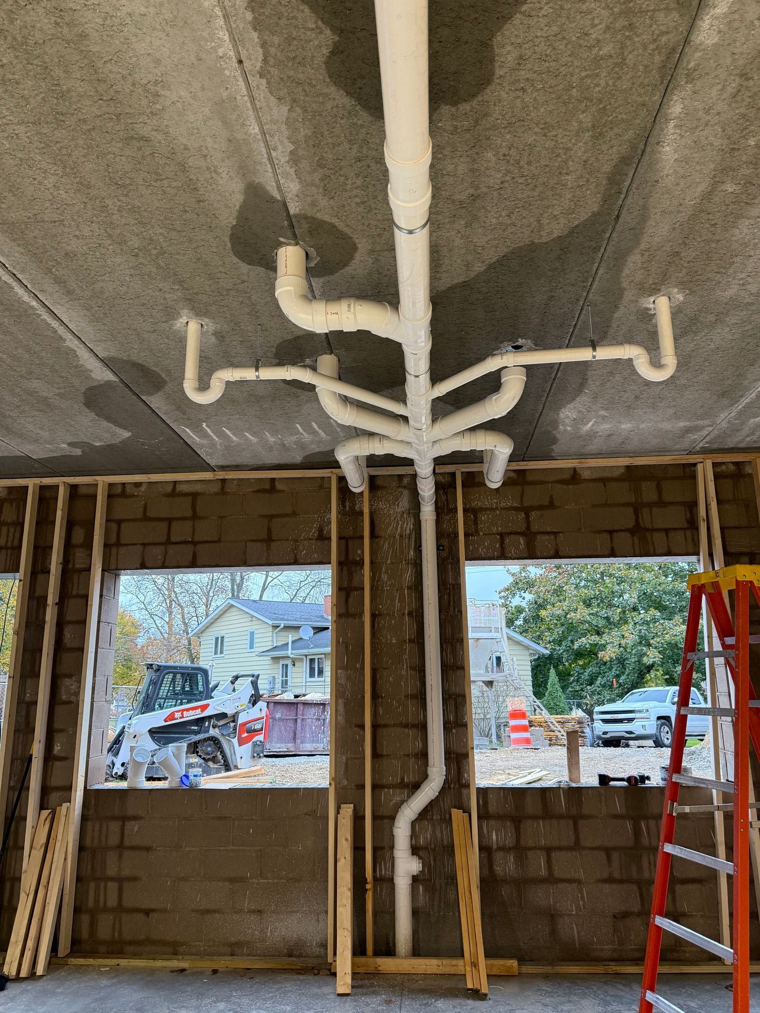 Interior construction site with exposed ceiling pipes, unfinished walls, and windows showing the street outside