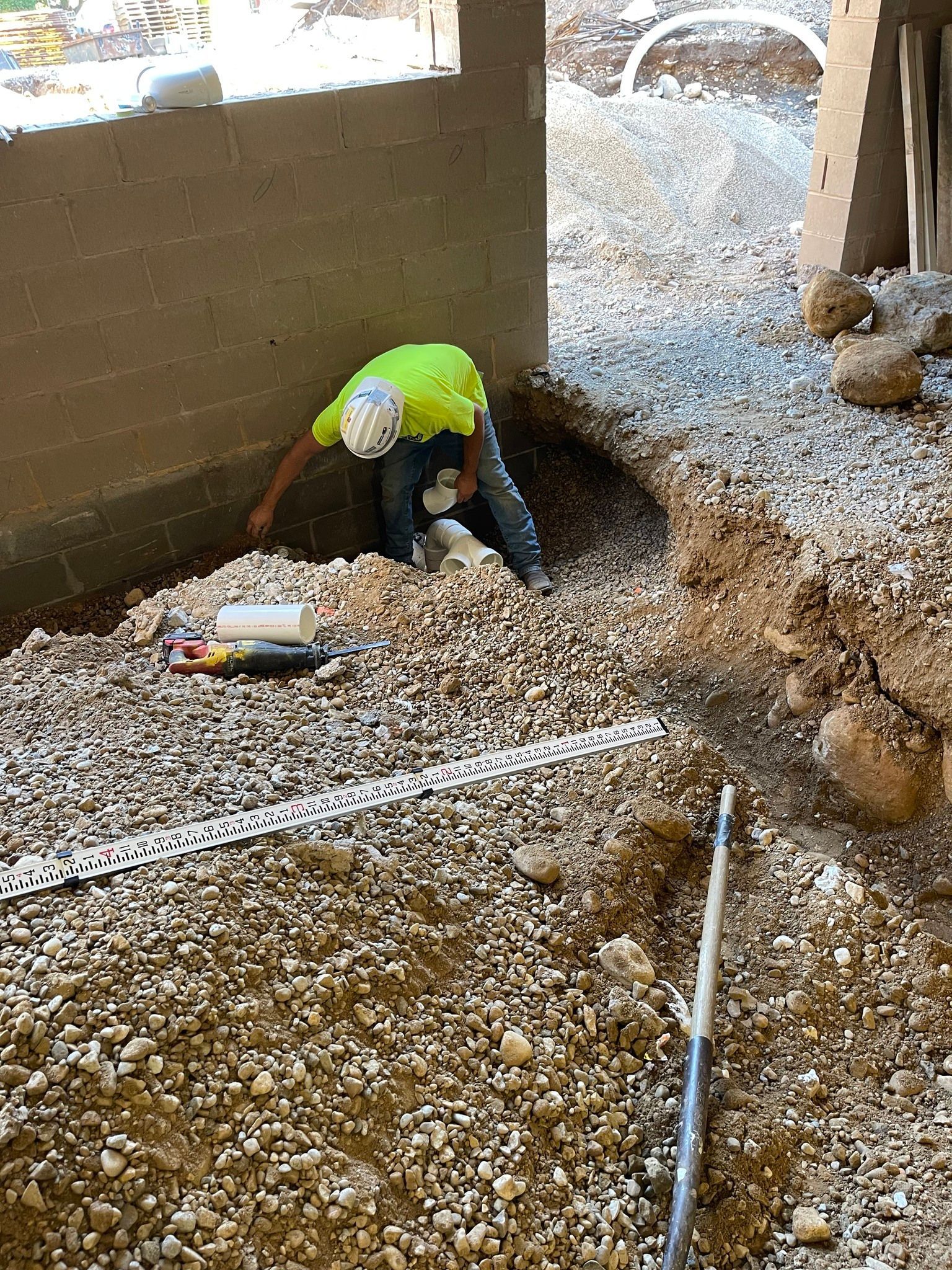 Worker in a neon shirt digging a hole in a gravel-covered construction site with tools nearby