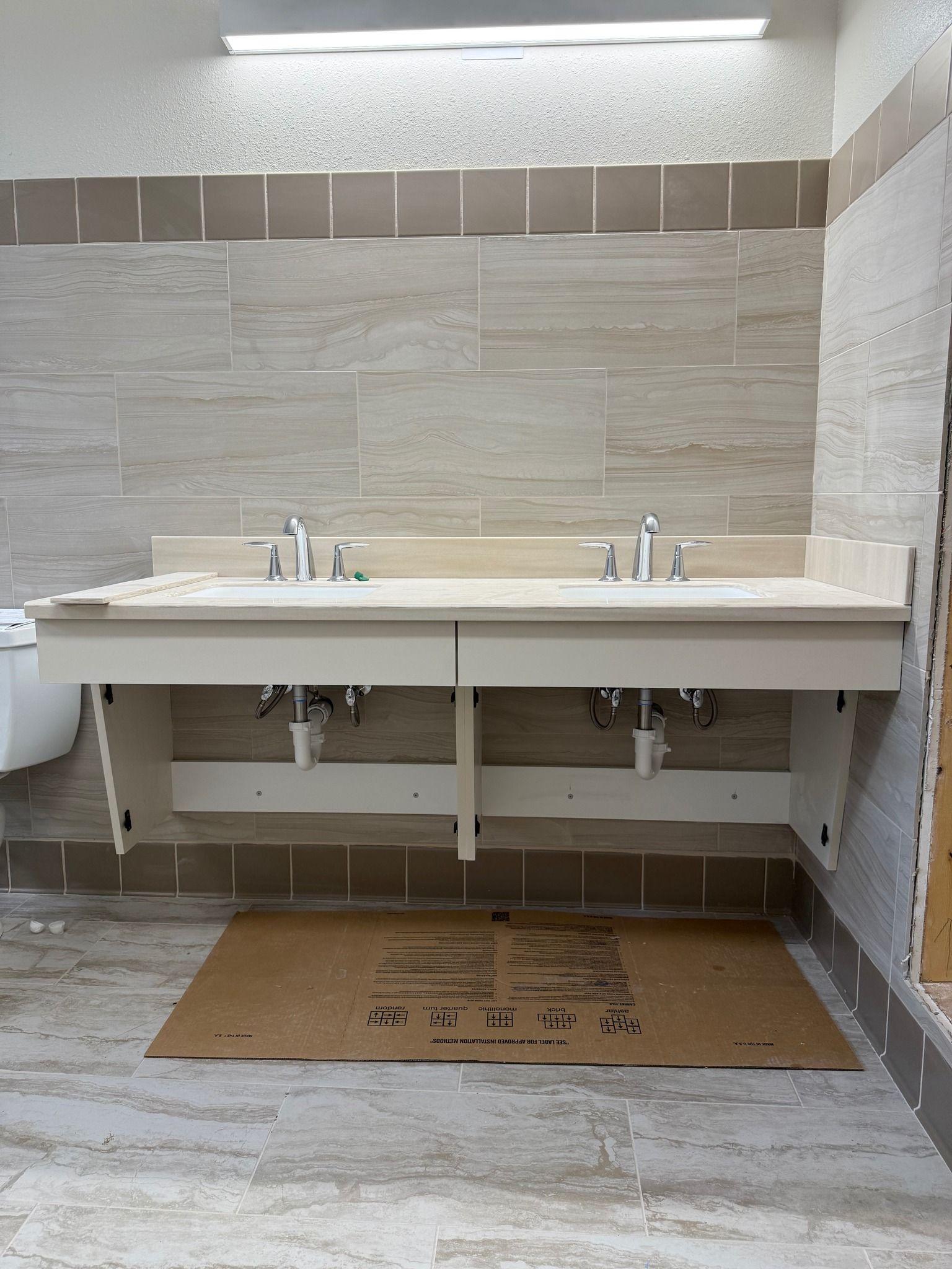 Public restroom with two white sinks, mirrors, and a tiled wall under bright lighting.
