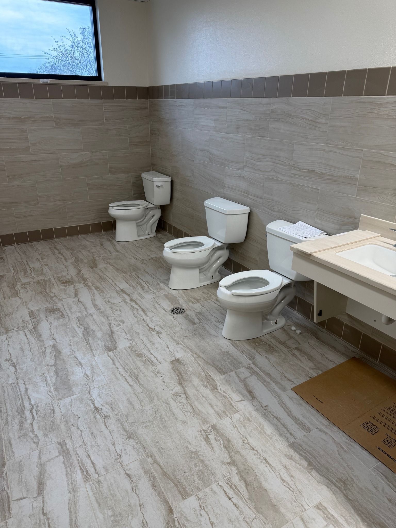 Accessible restroom with several toilets, beige tiled walls, and a window near the ceiling.