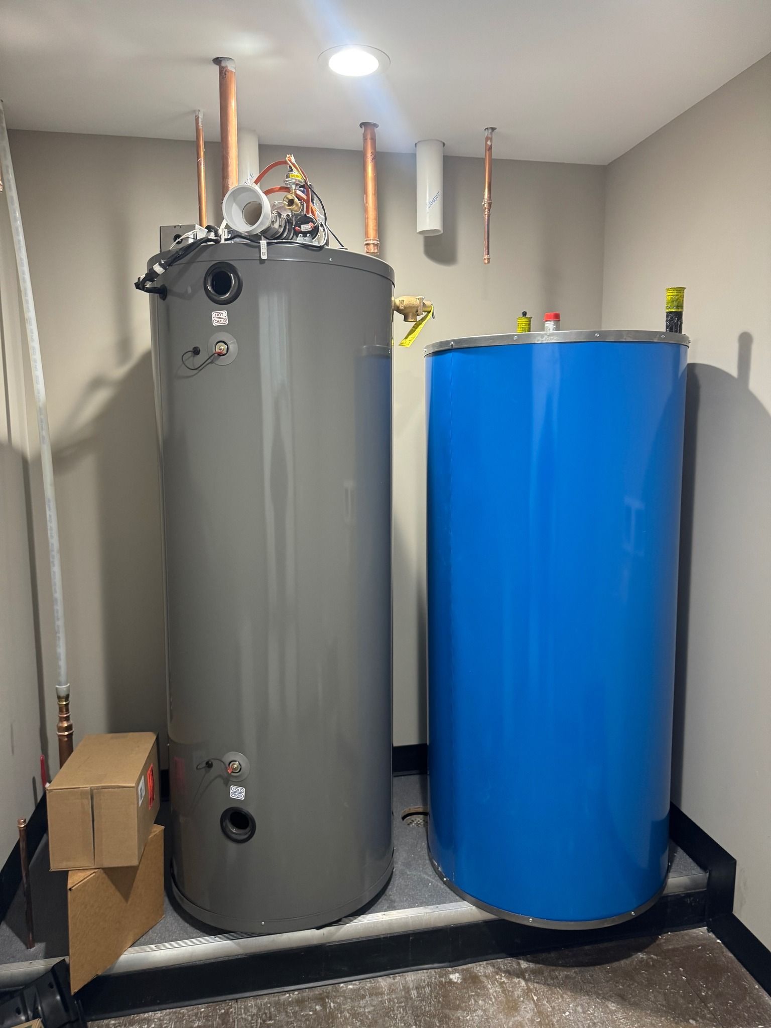 Two large water heaters, one gray and one blue, in a utility room with pipes above them
