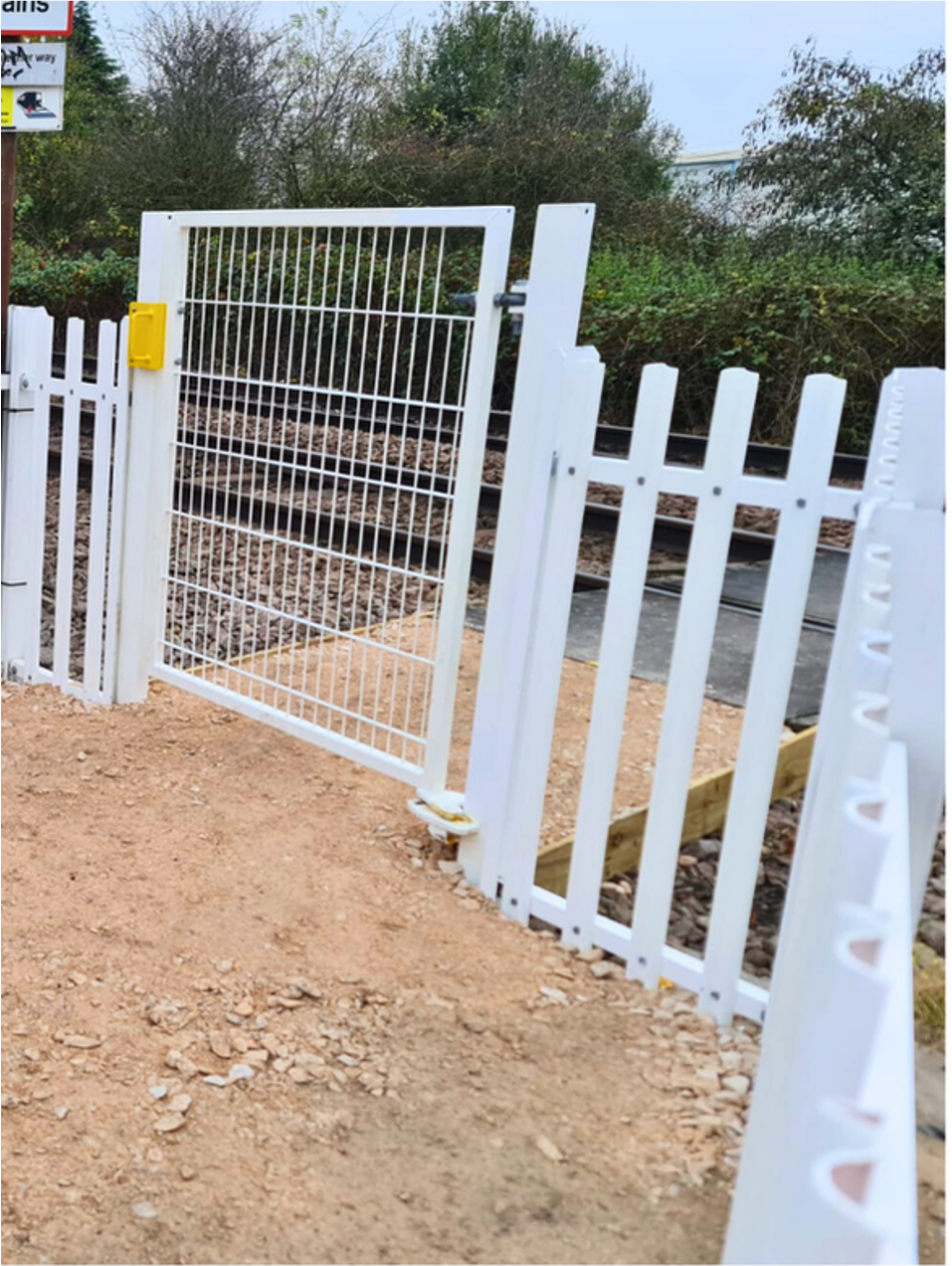 White fence and gate at a dirt path entrance beside green bushes under a clear sky