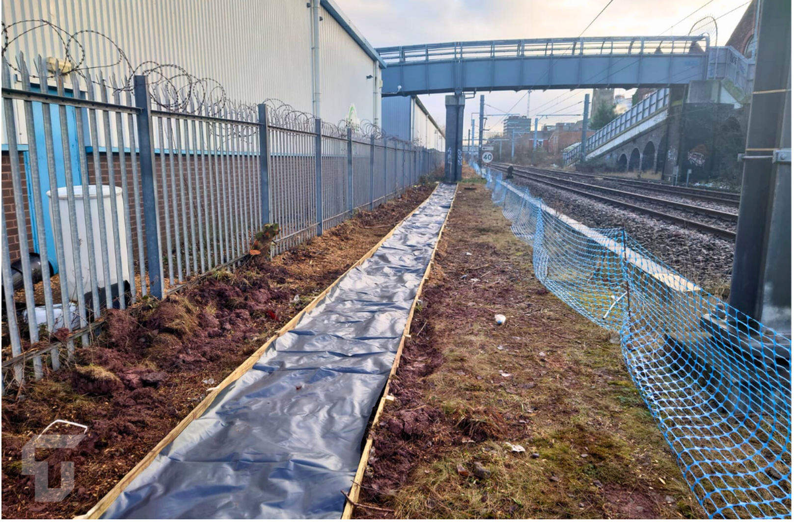 Railway side path with black sheeting, blue fencing, and train tracks under a pedestrian overpass