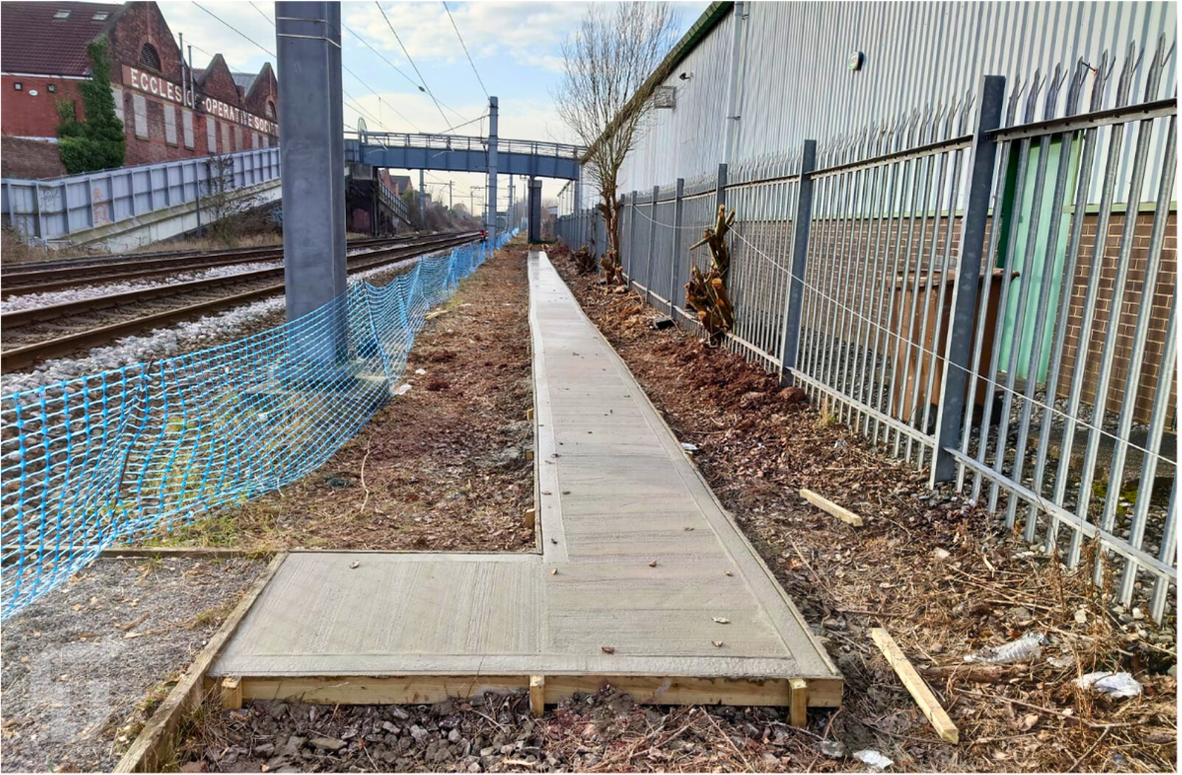 Narrow concrete path between blue fencing and a metal wall beside train tracks, with dry leaves and dirt.