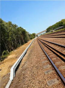 Railroad tracks curving through a wooded area under a clear blue sky, viewed at an angle.