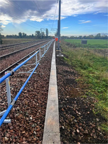 Railway tracks beside a concrete barrier and blue railing under a bright sky, with a person in red ahead.