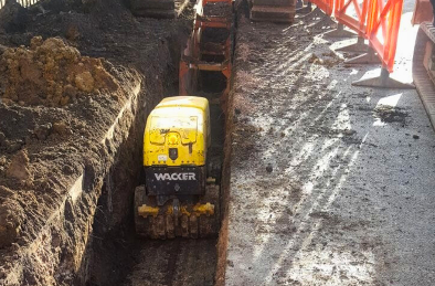 Yellow Wacker trench roller compacting soil in a muddy construction trench