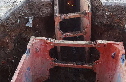Red excavator bucket trenching through wet soil beside a deep dug-out pit.
