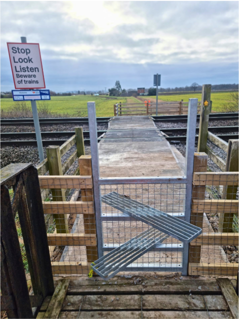 Wooden footbridge with a metal gate and a “Stop Look Listen” sign beside a field under cloudy skies