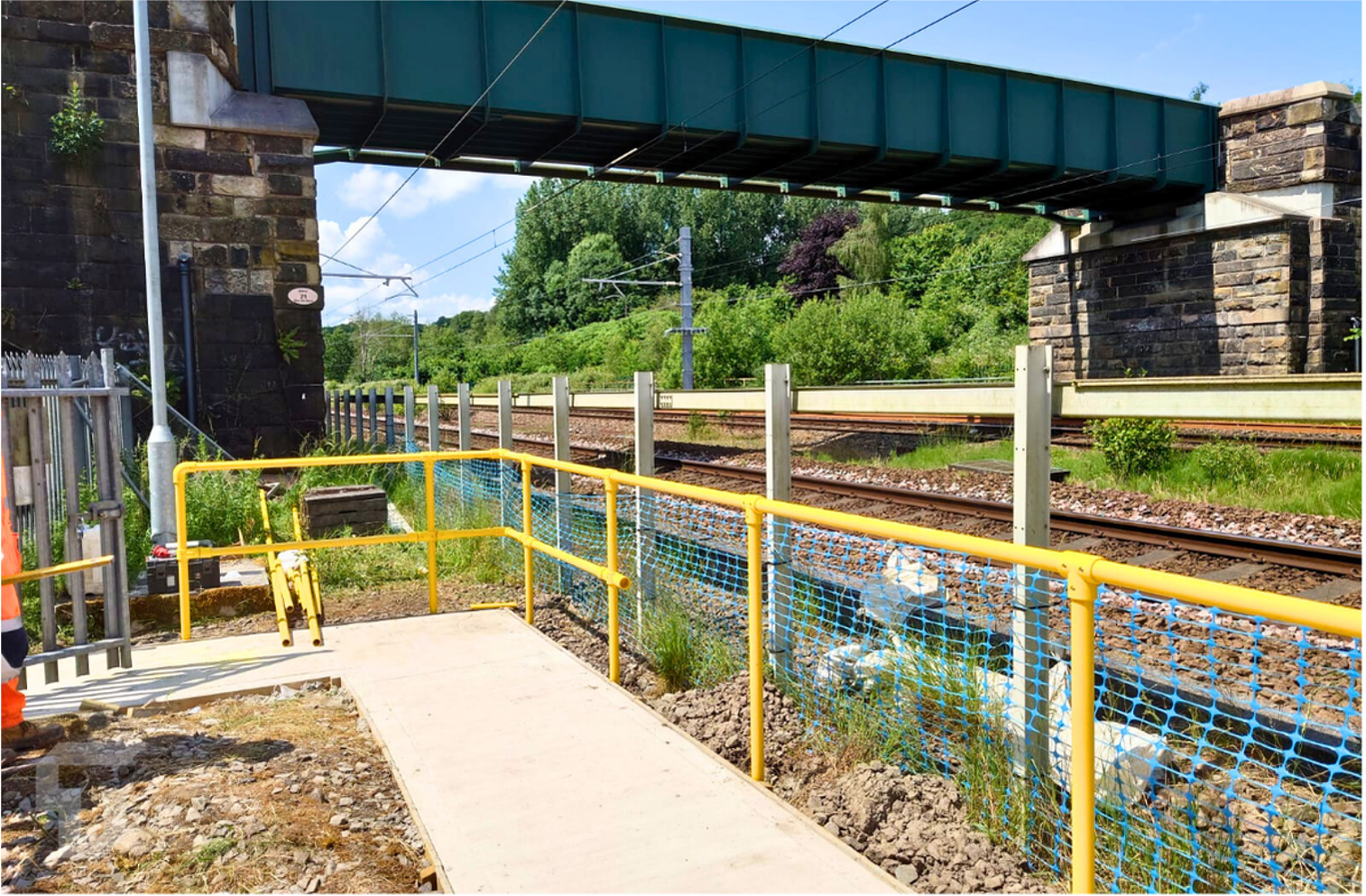 Yellow-railed walkway beside railroad tracks under a green bridge on a sunny day