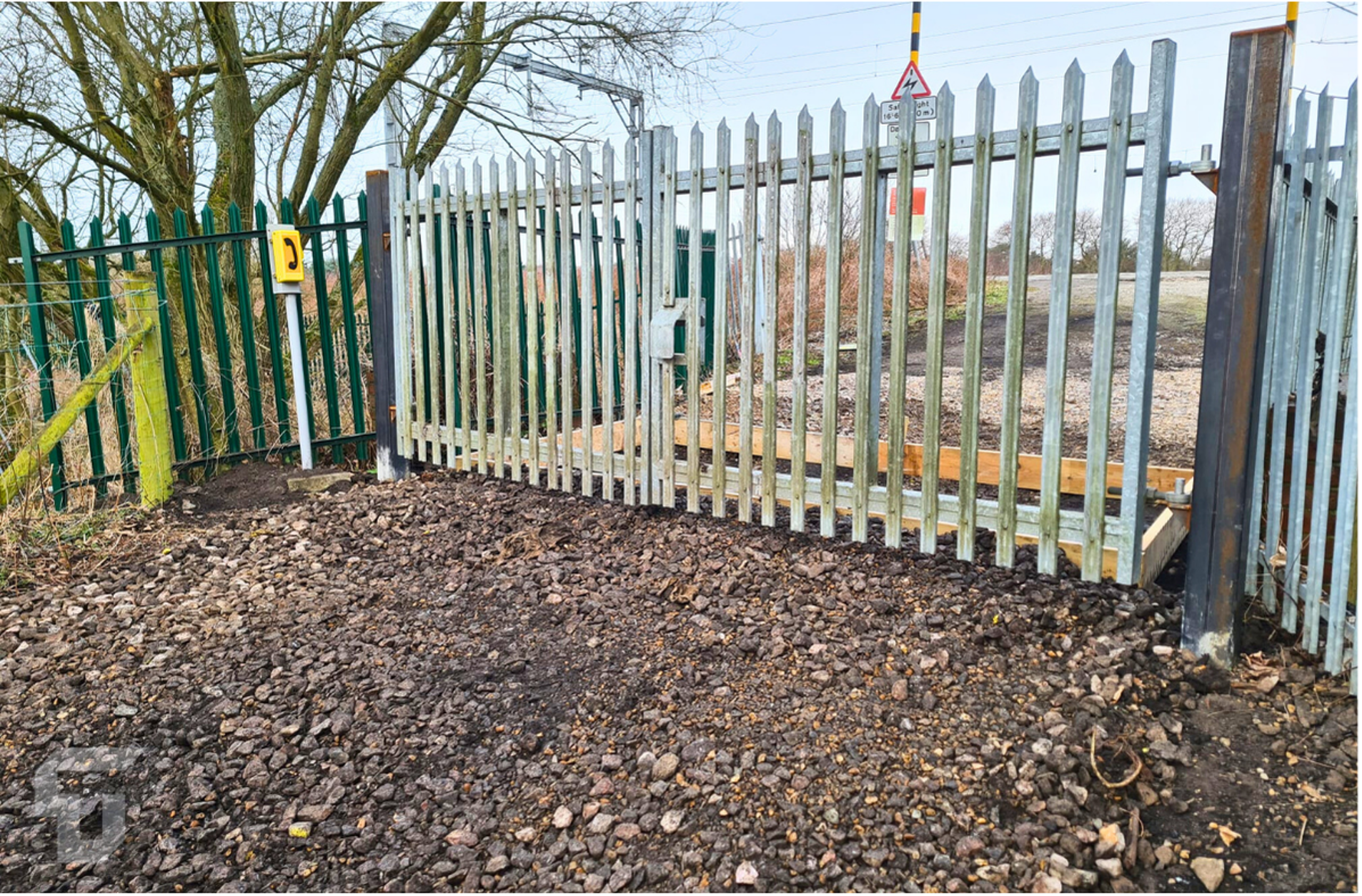 Metal fence and gate beside a gravel path, with a muddy lot and industrial site beyond.