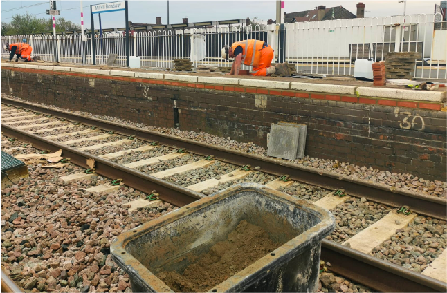 Railway tracks with a concrete work pit and workers in orange safety gear on a station platform