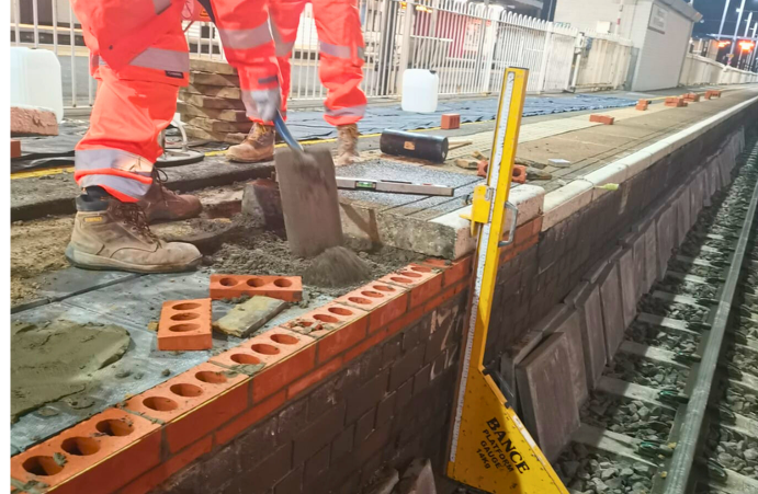 Workers in orange safety gear laying bricks along a waterfront construction edge