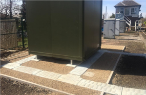 Large green utility shed on a paved base beside a driveway, with a house in the background.