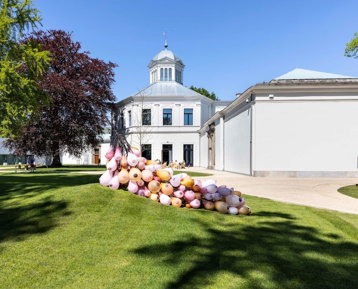 Een sculptuur, bestaande uit bleke, ballonachtige vormen, staat op een grasveld voor een wit gebouw met een centrale koepel.