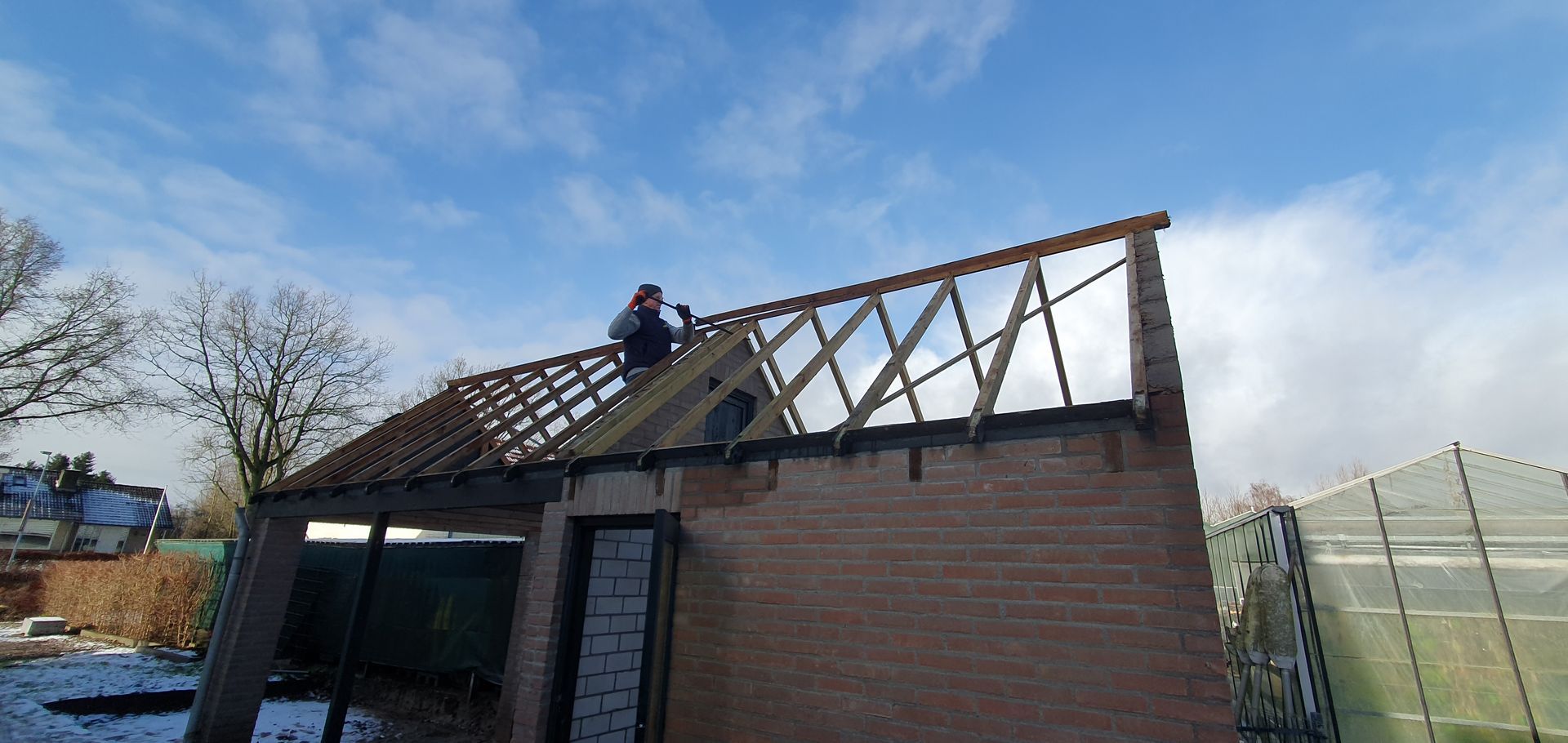 Een persoon werkt op het dak van een bakstenen gebouw, met een blauwe lucht erboven, bomen op de achtergrond en een kas.