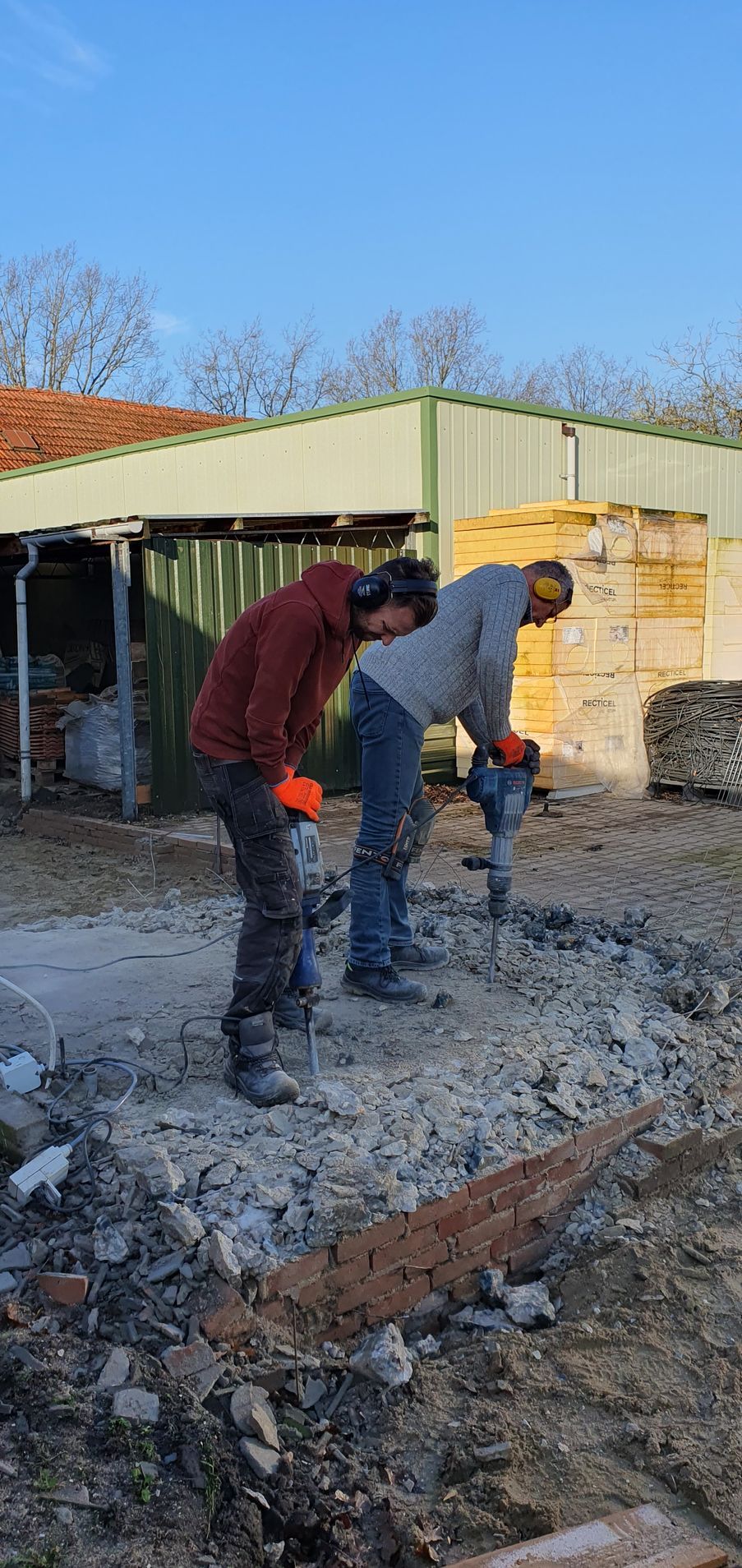 Twee mensen gebruiken drilhamers op een betonnen oppervlak buiten een gebouw op een zonnige dag.