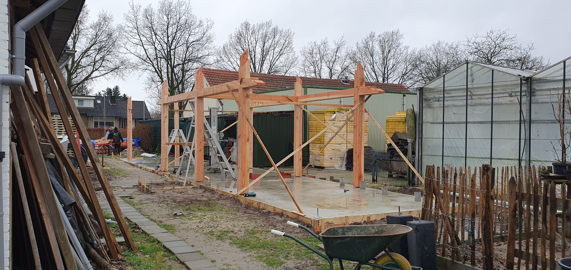 De bouw van een houten frameconstructie op een erf, met op de achtergrond een gedeeltelijk gebouwd gebouw.