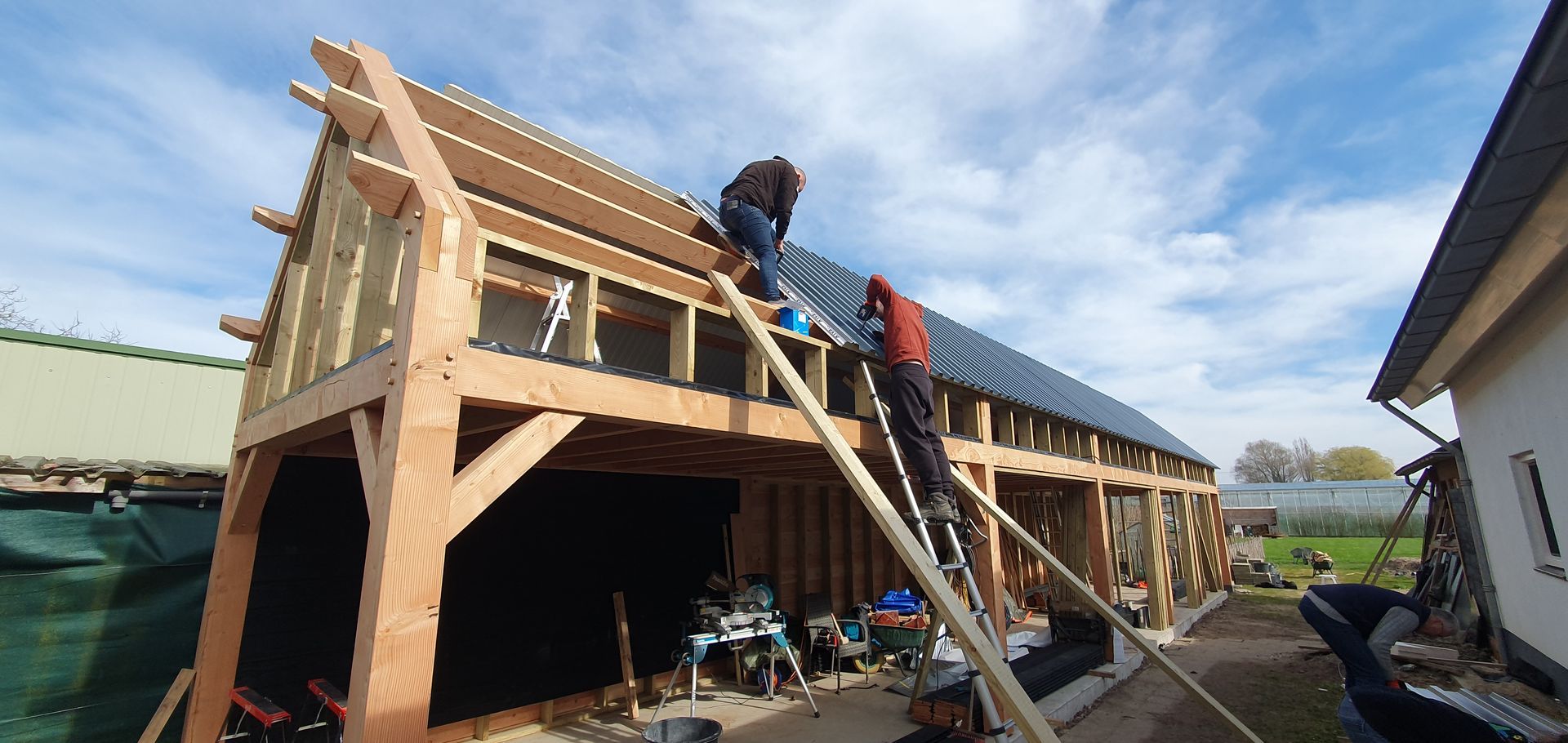 Twee mensen zijn bezig met het aanbrengen van dakbedekking op een houten gebouwframe. Buiten, zonnig weer.