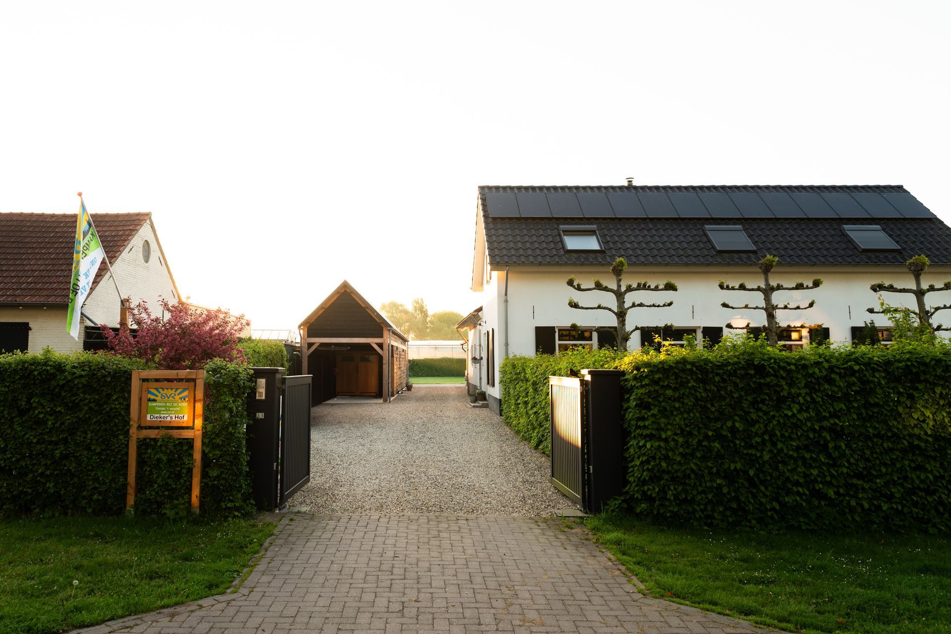 Witte boerderij met carport, oprit en heggen, verlicht door de zonsondergang.