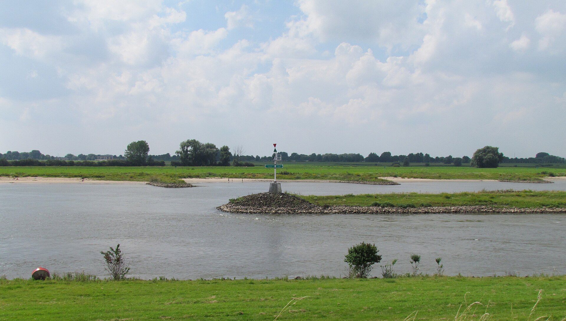 Een met stenen beklede golfbreker steekt uit in een kalme rivier onder een bewolkte blauwe hemel, met grasrijke oevers en bomen in de verte.