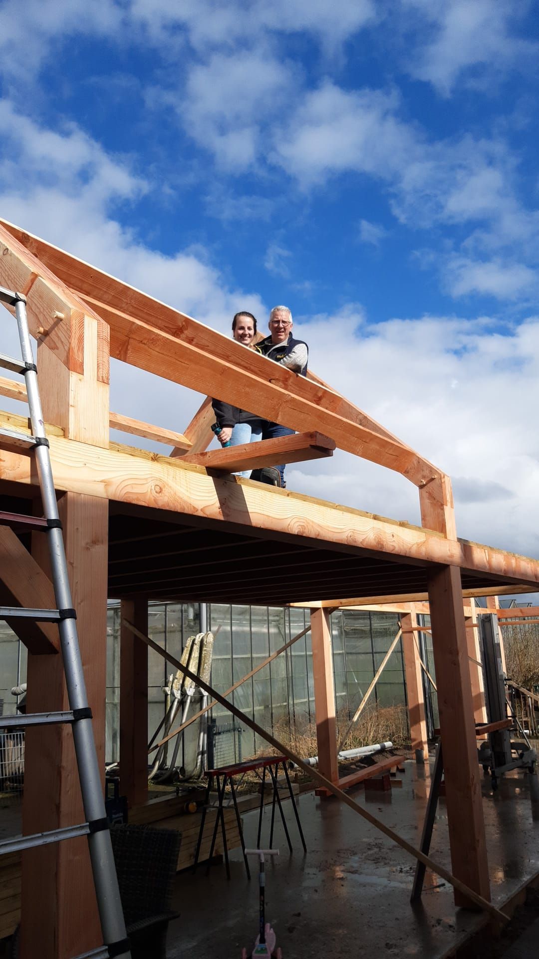 Twee mensen staan ​​op een houten constructie in aanbouw, tegen een gedeeltelijk bewolkte hemel.