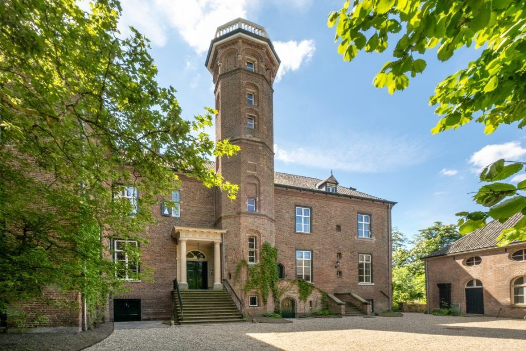 Een statig bakstenen landhuis met een centrale toren en stenen toegangstrappen, omgeven door groene bomen op een zonnige dag.