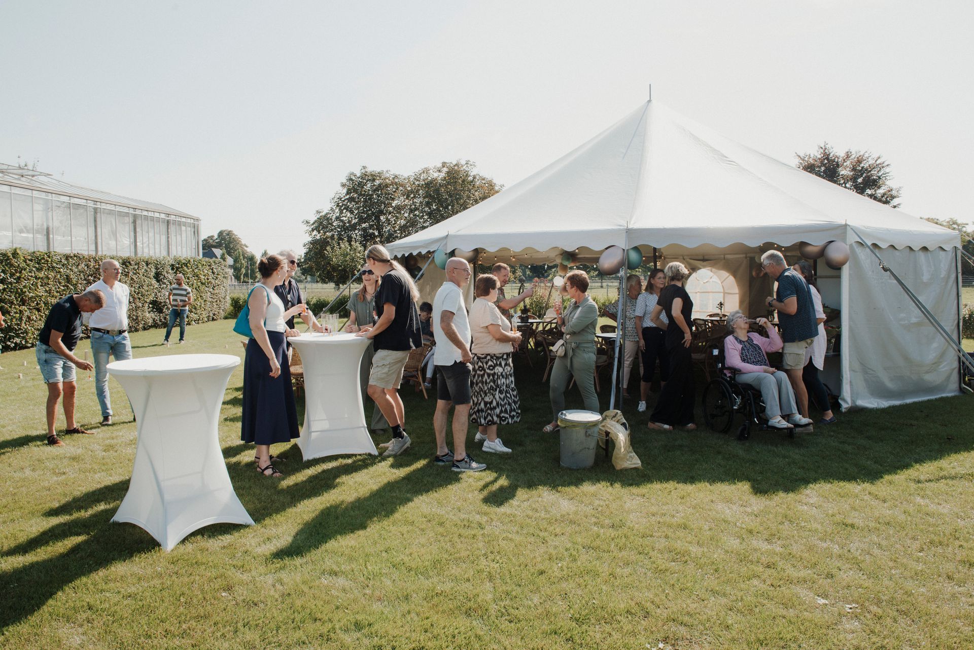 Mensen verzamelen zich bij een evenement in een tent in de openlucht; sommigen staan ​​aan hoge tafels, anderen praten in de buurt van een gehandicapte persoon in een rolstoel.