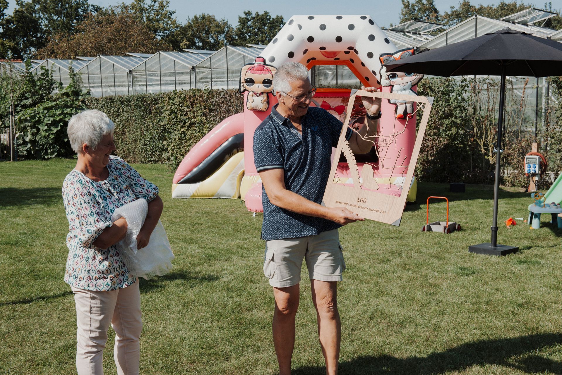 Een man houdt een roze kartonnen figuur vast, een vrouw kijkt toe, in een achtertuin met een springkasteel.