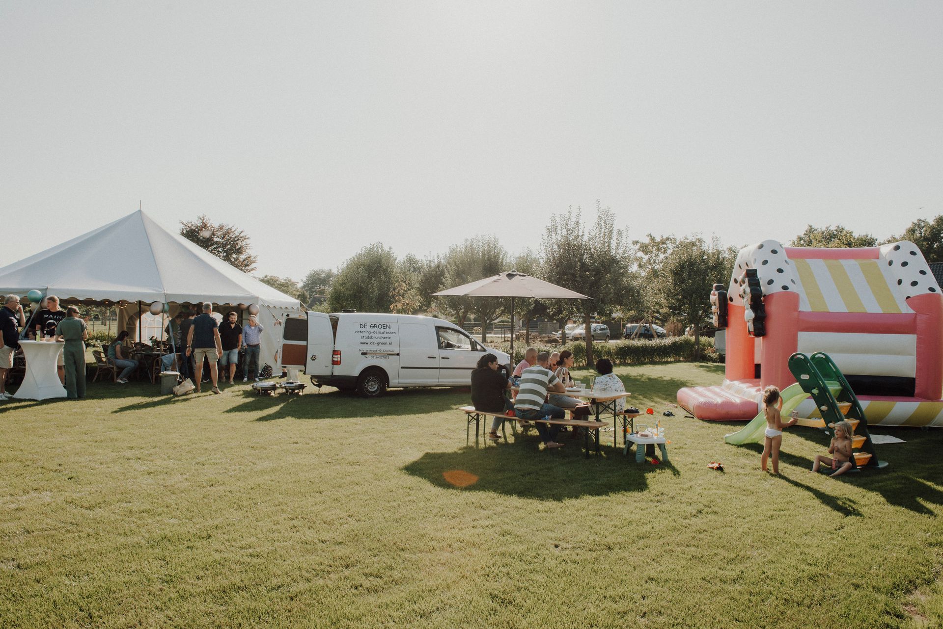Een buitenfeest met een tent, springkussen, foodtruck en mensen die aan een picknicktafel zitten.