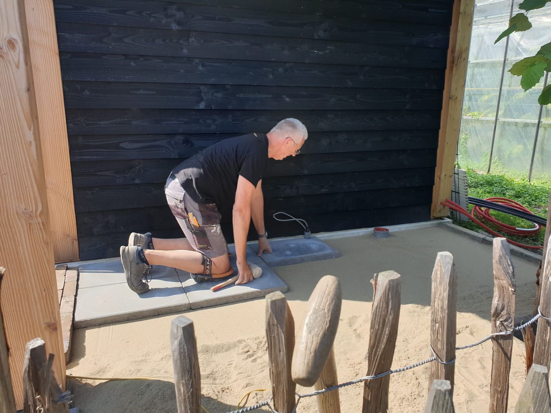 Een man knielt en legt straatstenen op een zanderige ondergrond naast een donkere houten muur en schutting.
