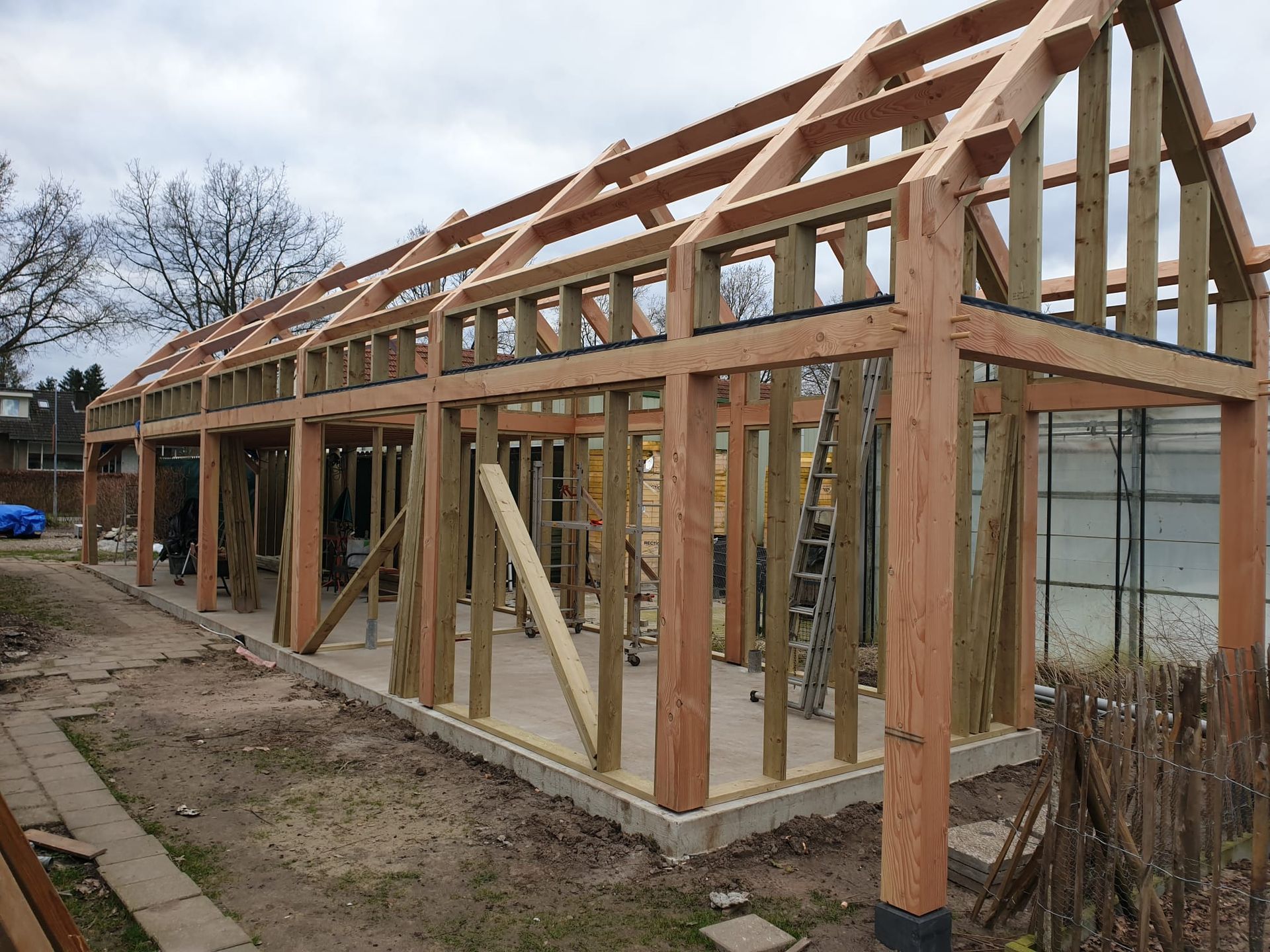 Houten frame in aanbouw, buiten op een betonnen fundering, met een zichtbare ladder.