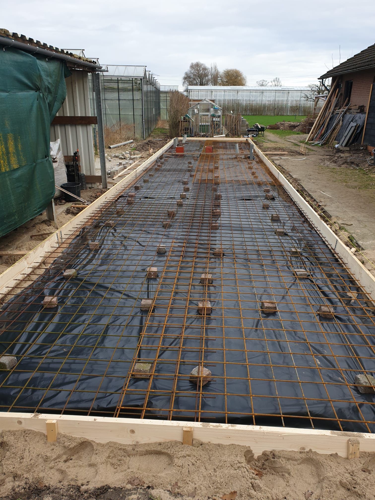 Bouwplaats: rechthoekig houten frame, wapeningsnet, zwart zeil, bakstenen en omliggende gebouwen.