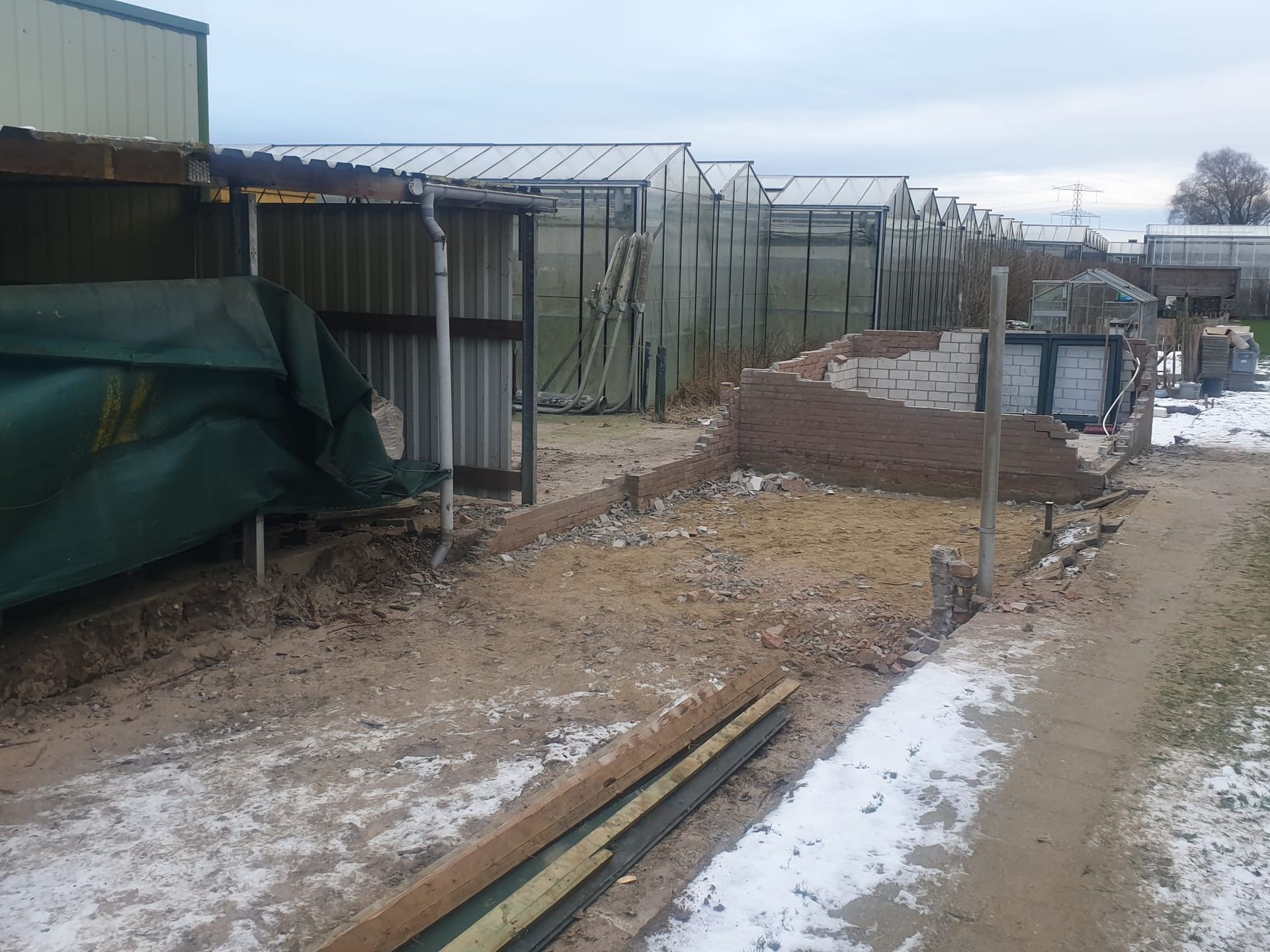Een zandpad, overblijfselen van een bakstenen gebouw, metalen schuren, kassen op de achtergrond, een winterlandschap met sneeuw.