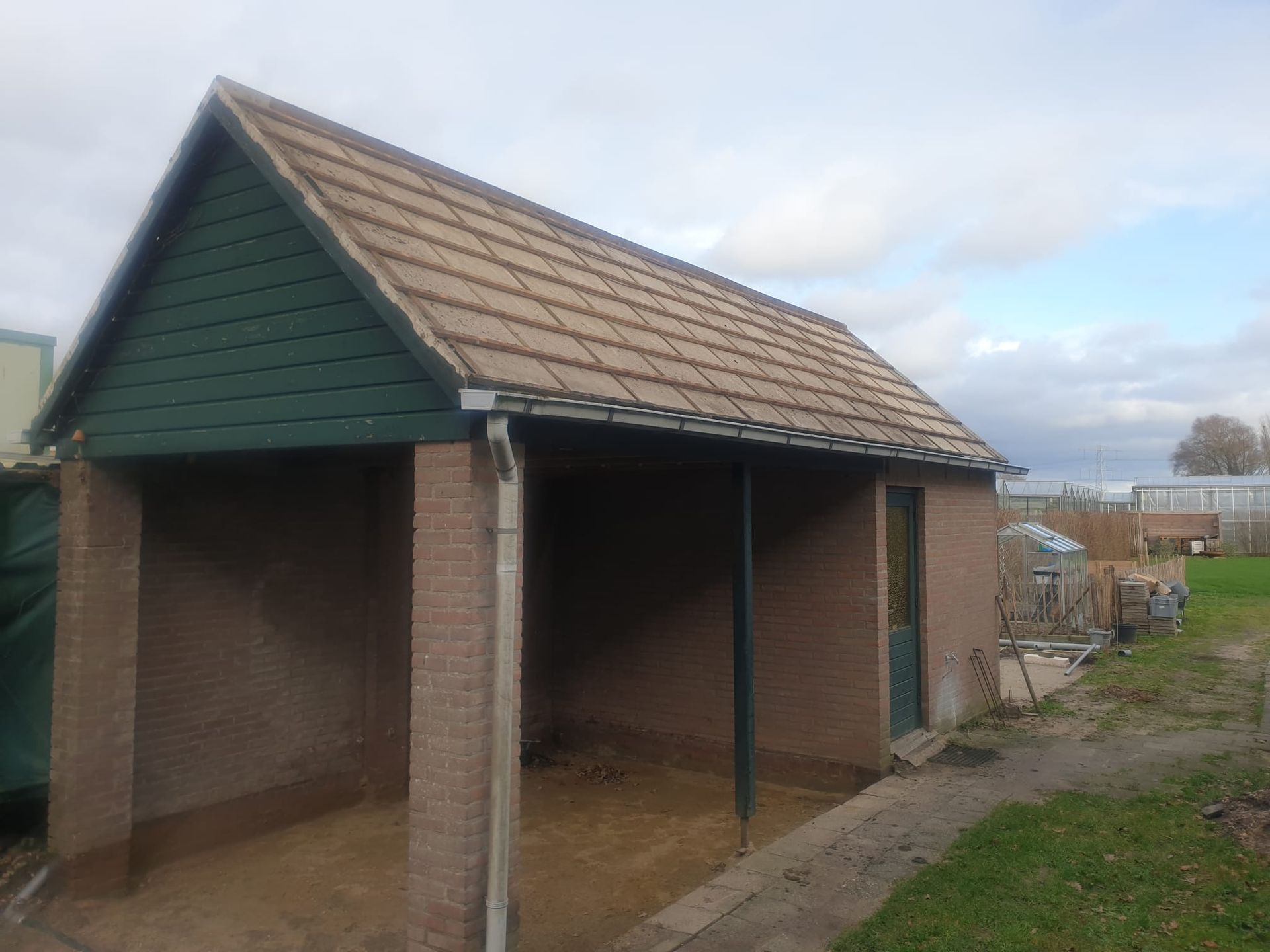 Een bakstenen schuur met bruine dakpannen en een groene gevel tegen een bewolkte hemel.