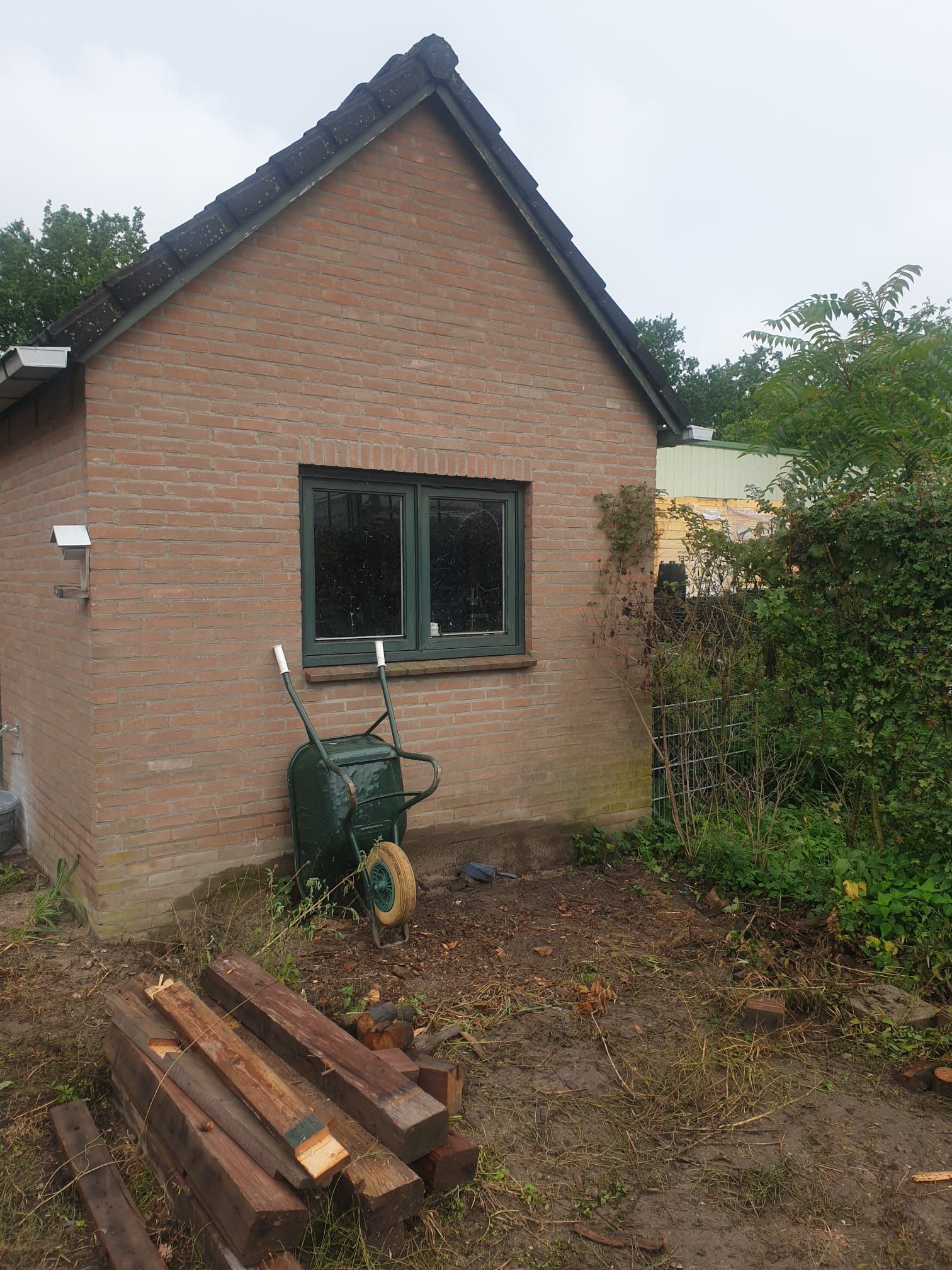 Een bakstenen gebouw met een raam met groene kozijnen. Buiten staat een kruiwagen. Op de grond ligt een stapel hout.