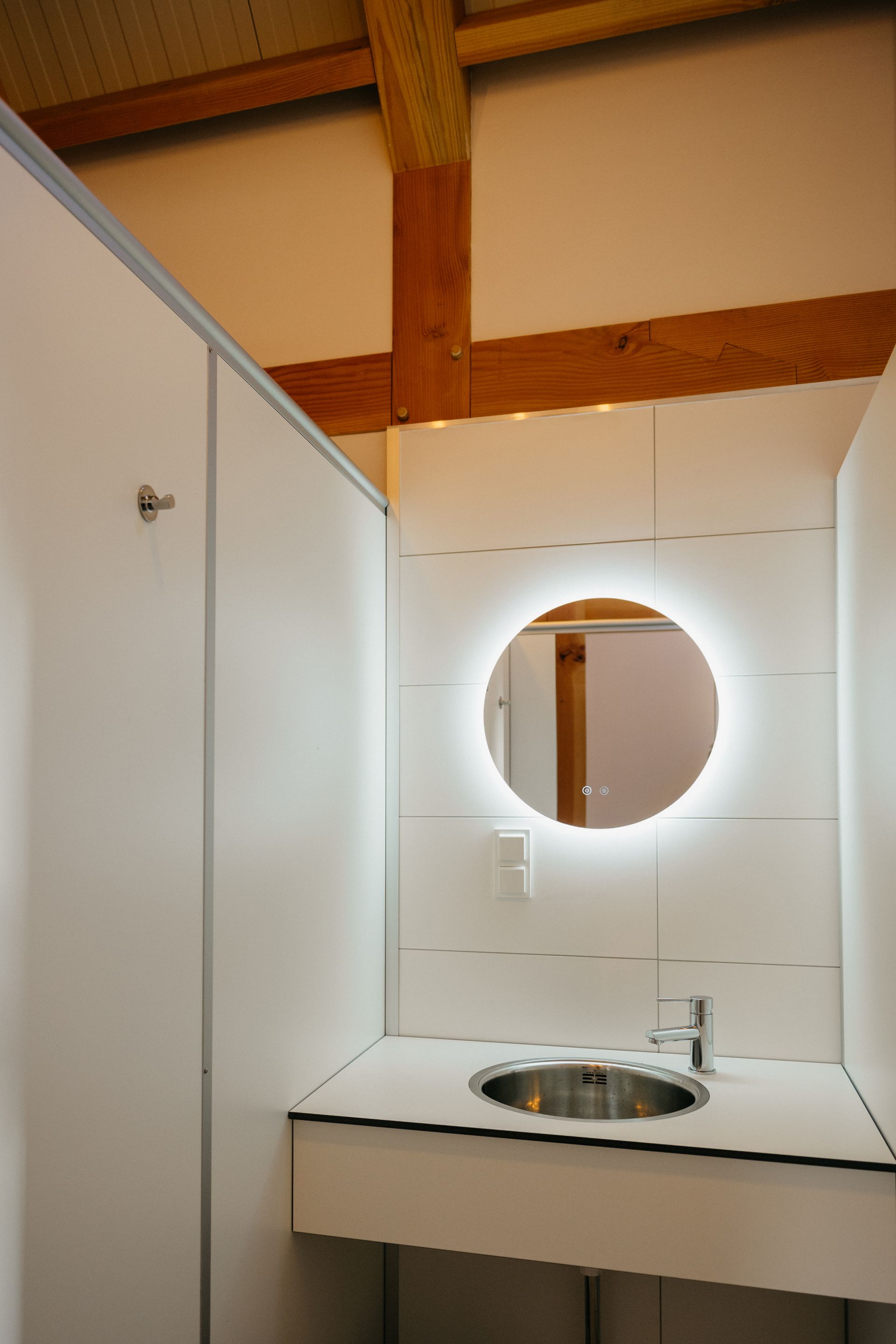 Witte toiletcabine met een ronde, verlichte spiegel boven de wastafel. Houten balken in het plafond.