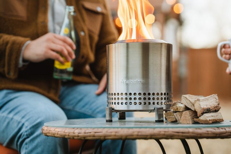 Een Solo Stove vuurkorf met vlammen, een persoon die een drankje vasthoudt en brandhout op een tafel.