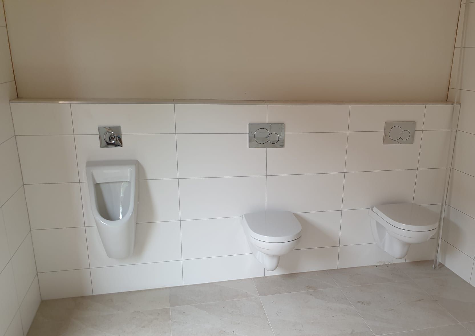 Badkamerwand met witte tegels, een urinoir, twee aan de muur gemonteerde toiletten en spoelknoppen.