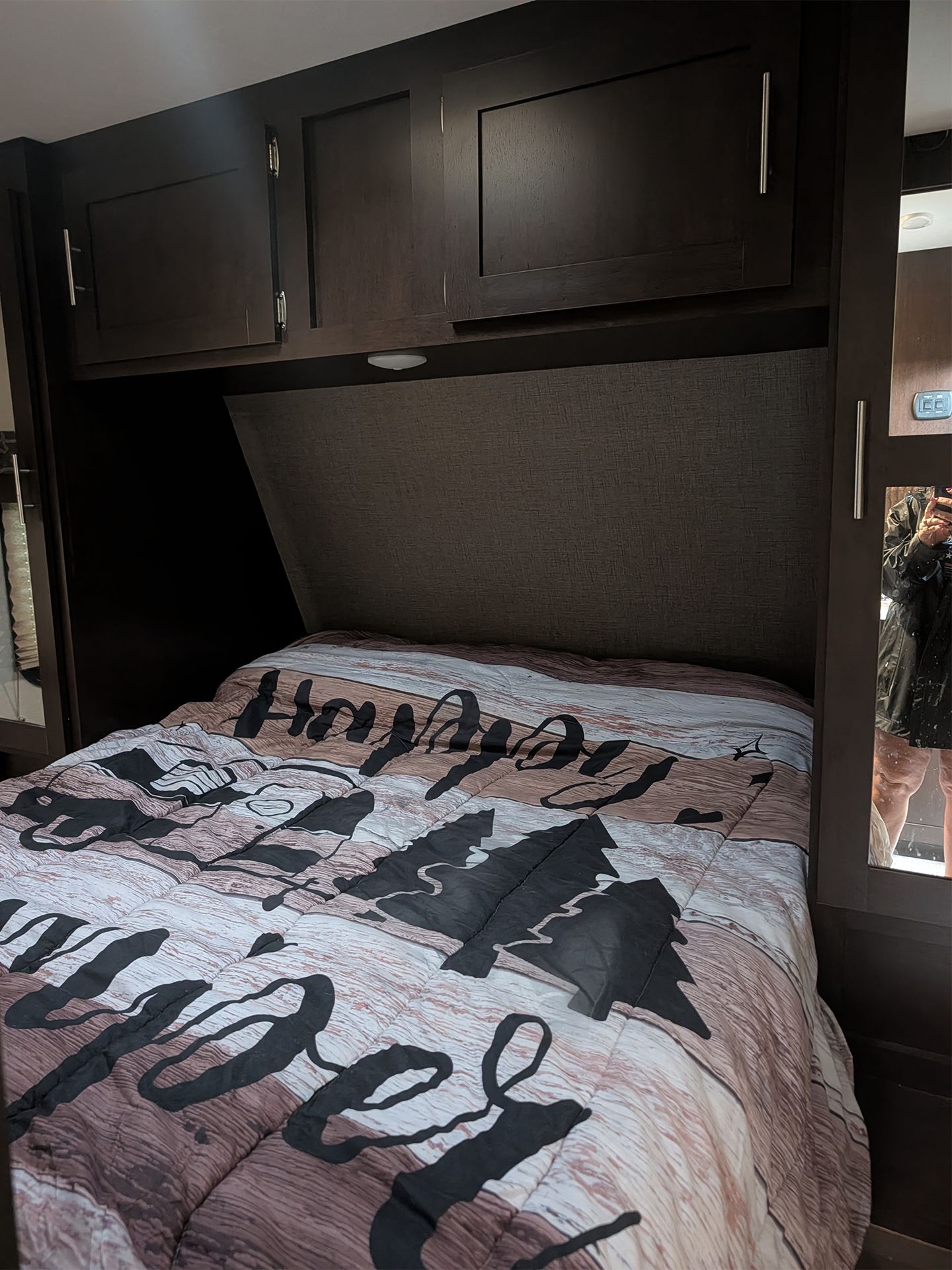 Bedroom with a bed made up in a gray and black tree-patterned blanket beneath dark overhead cabinets.