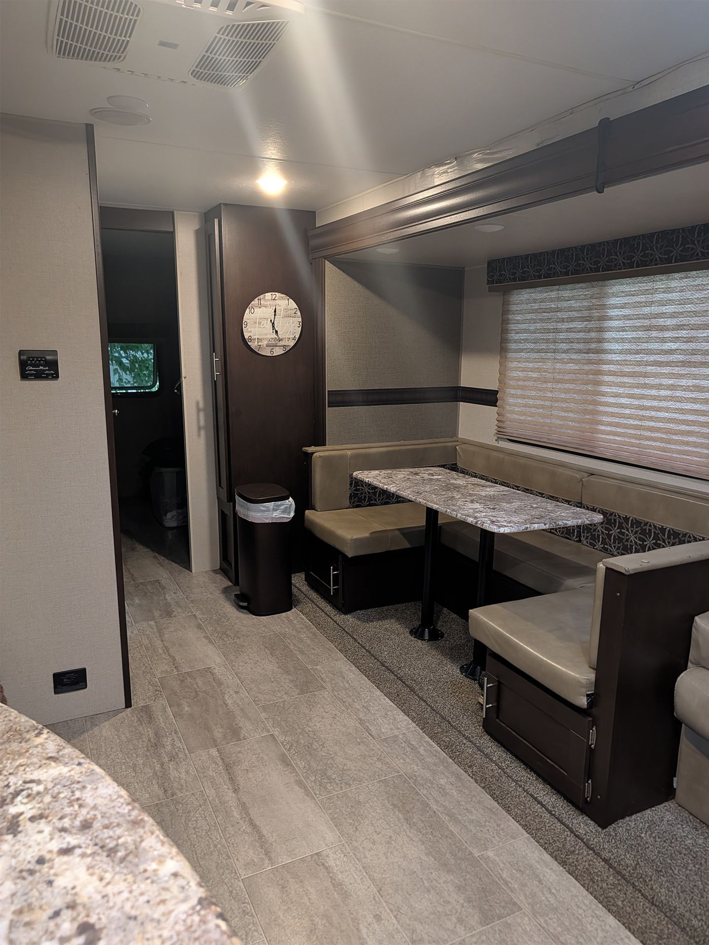 Compact RV dining area with booth seating, table, window blinds, and clock along a narrow walkway.