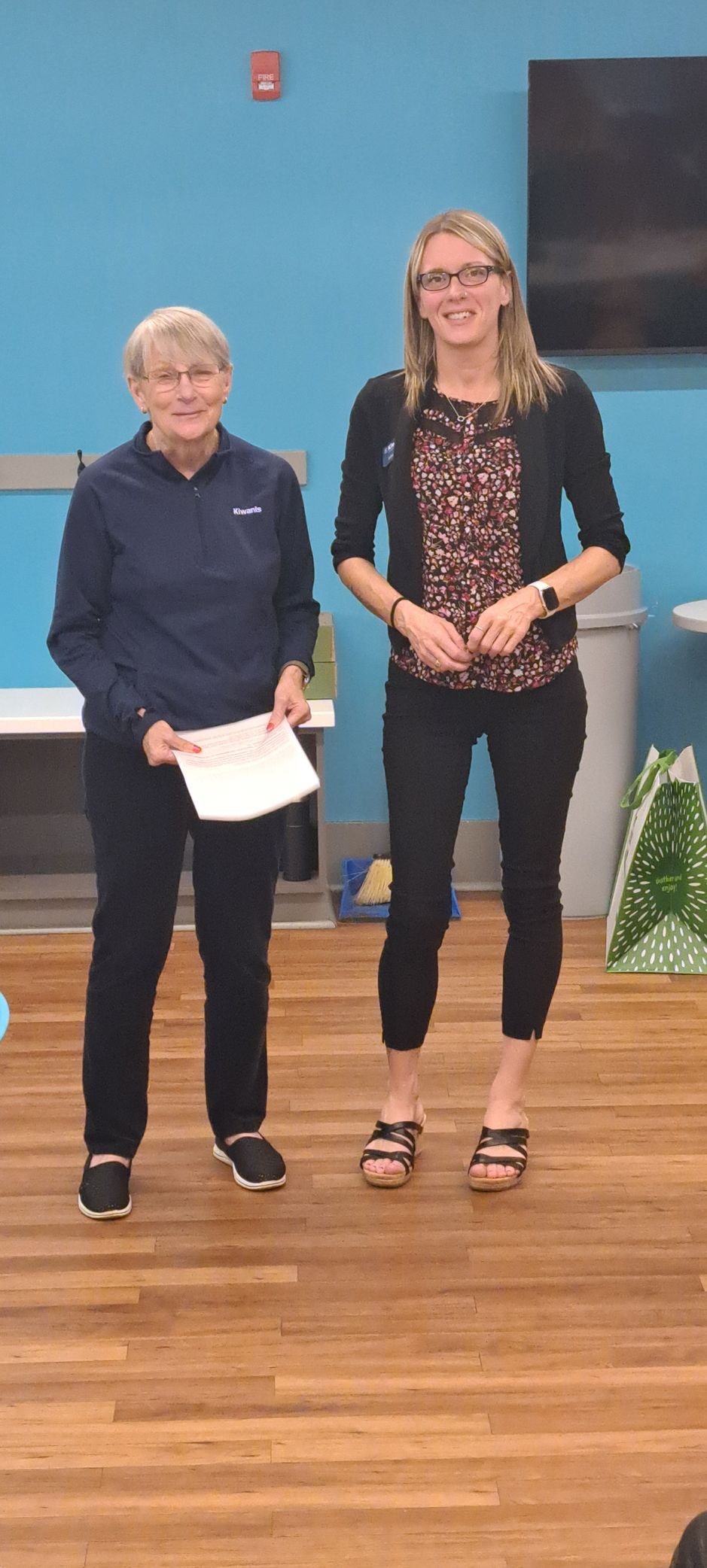 Two women stand indoors in front of a blue wall and a wooden floor. One holds a paper.